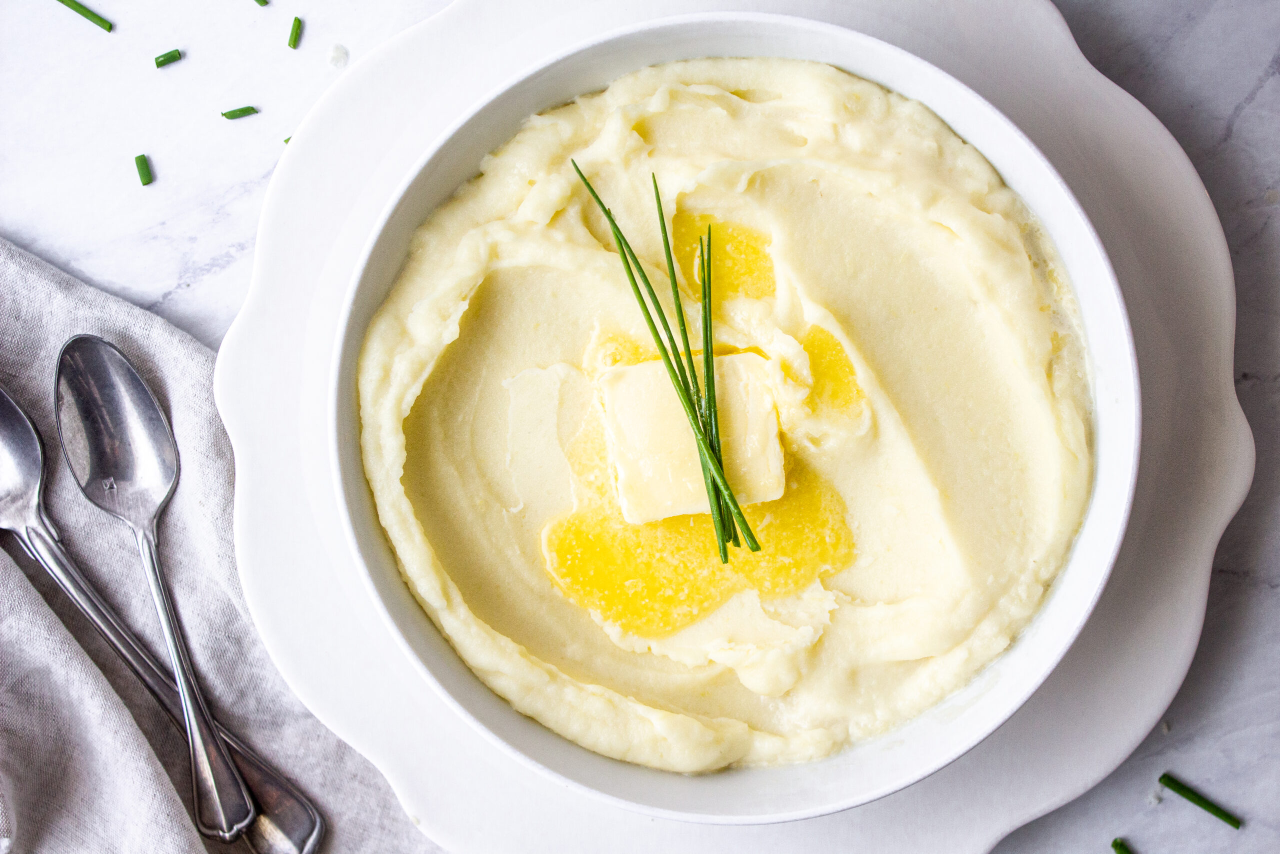 mashed potatoes in a white bowl on top of a scalloped white plate and melted butter and chive sprigs