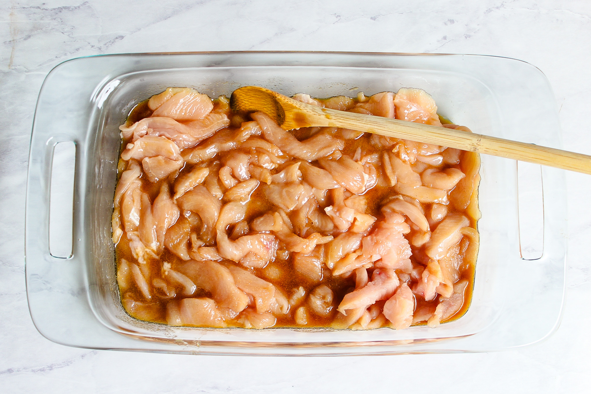 Chinese chicken marinating in a glass baking dish with wooden spoon