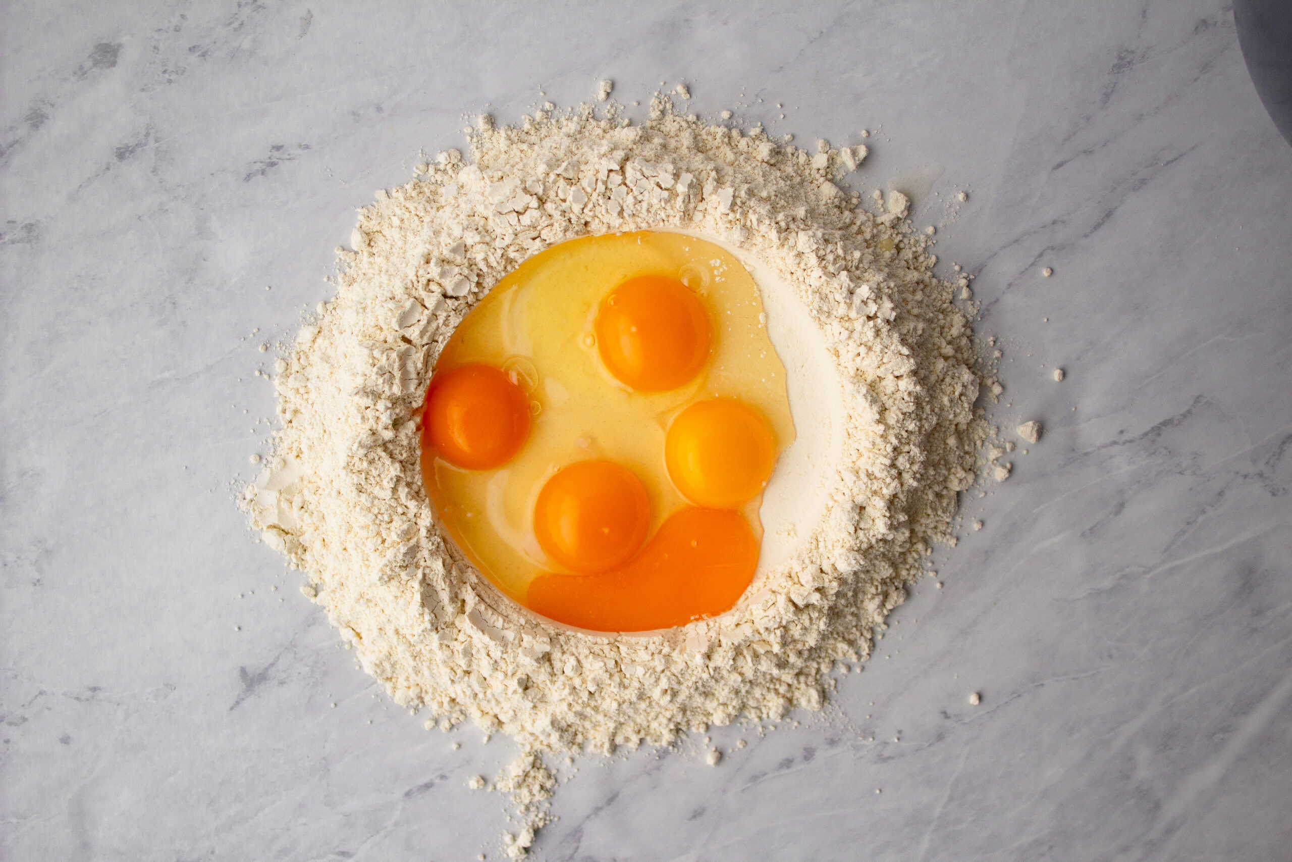 Eggs and egg yolks cracked into a well of einkorn flour on a marble surface, the first step in making fresh einkorn pasta dough
