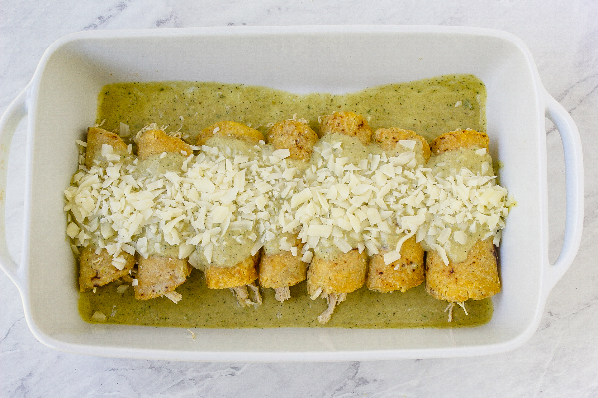 enchiladas in a single row in a baking dish with salsa verde poured on top and around topped with cheese before baking