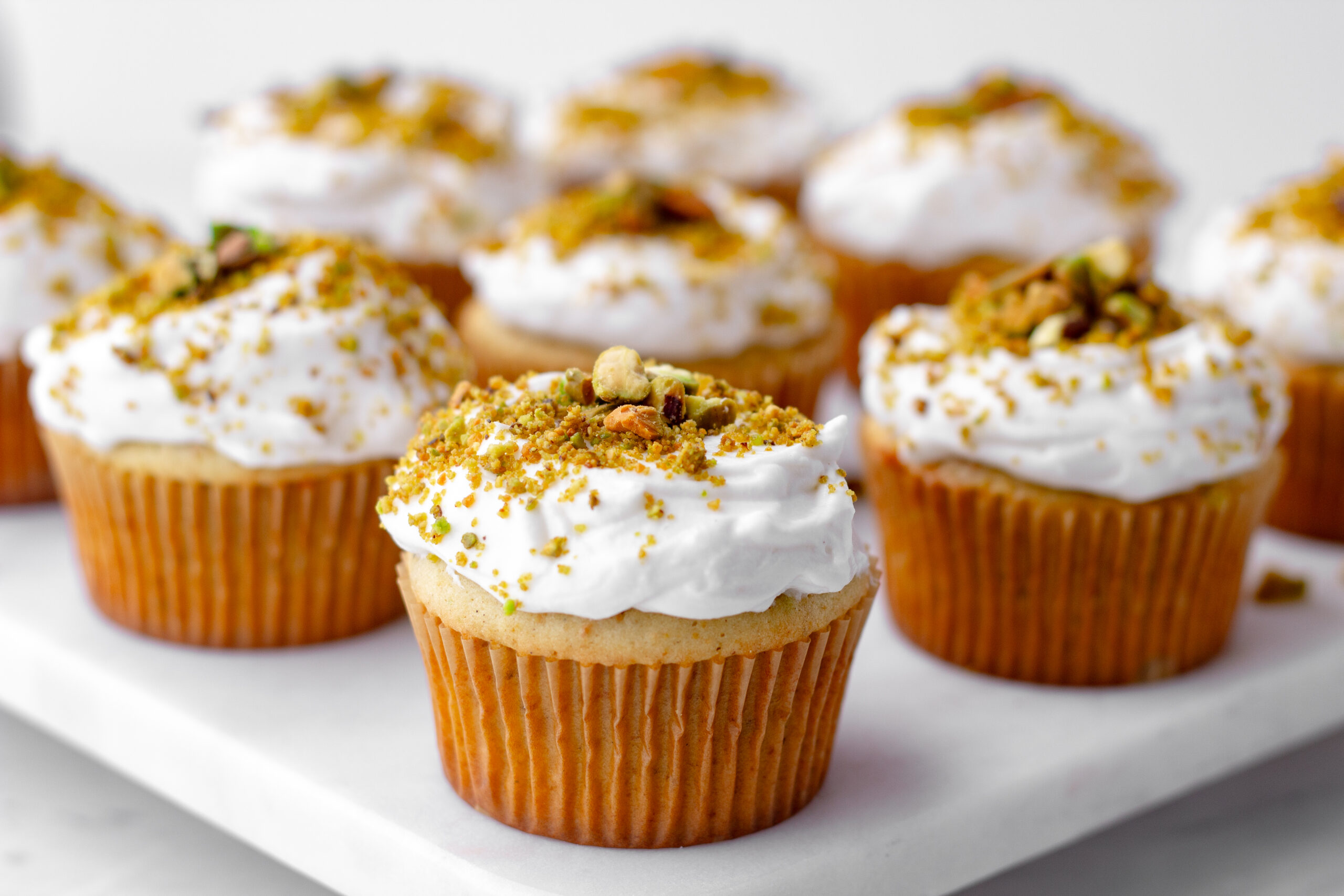 einkorn cupcakes with white frosting and pistachios lined up in a diagonal formation on a whit marble board