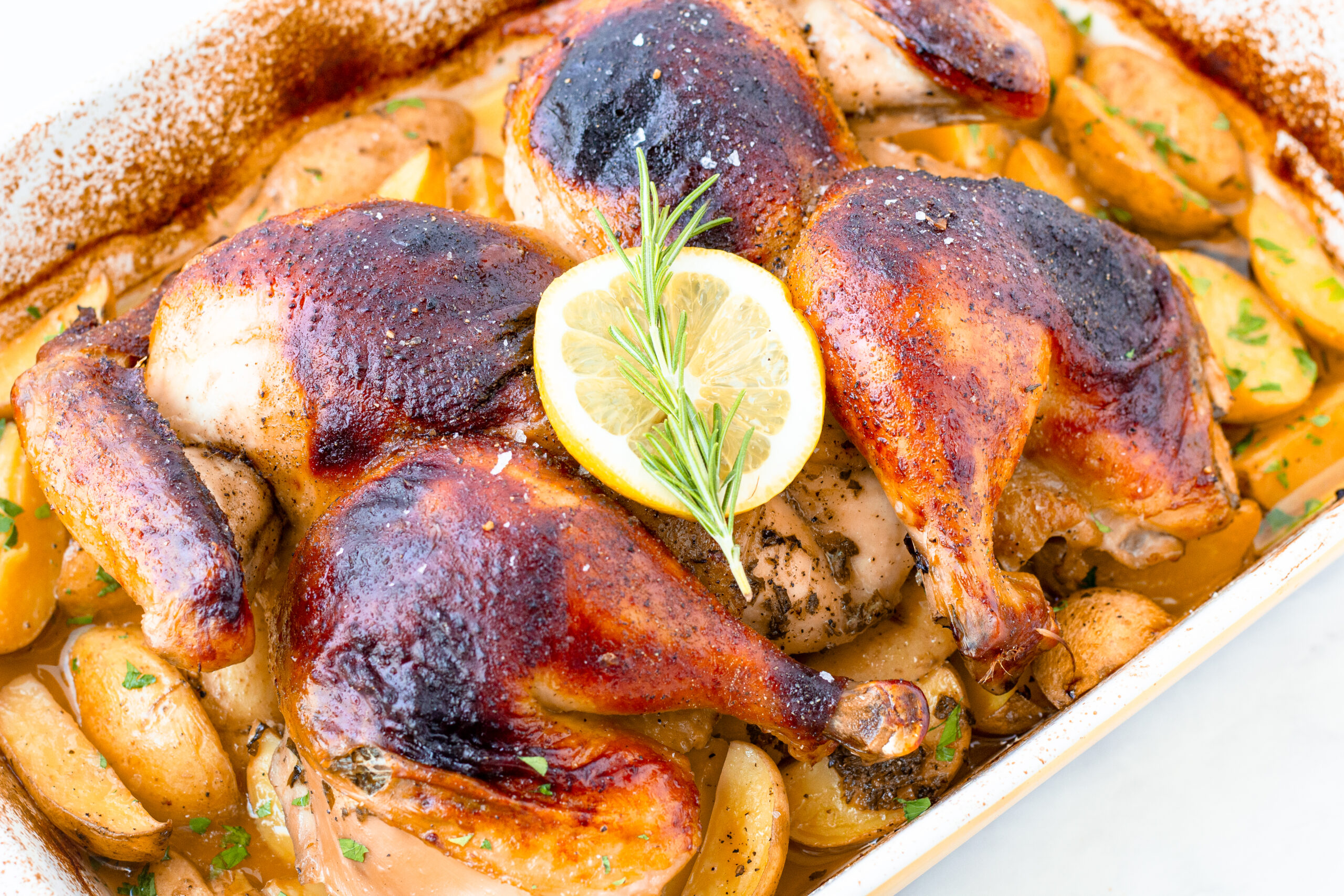 golden spatchcock chicken in roasting pan with gold potatoes and a lemon slice and rosemary sprig