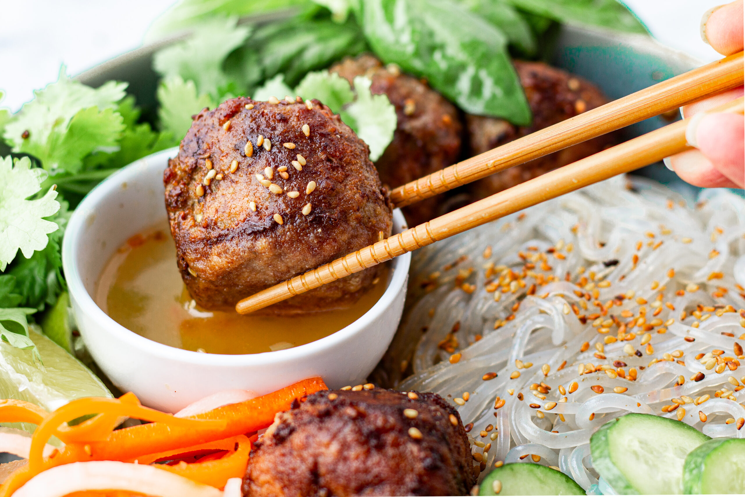chopsticks holding a vietnamese charred meatballs in ablue bowl with sauce pickled carrots and daikon basil cilantro cold noodles ans sesame
