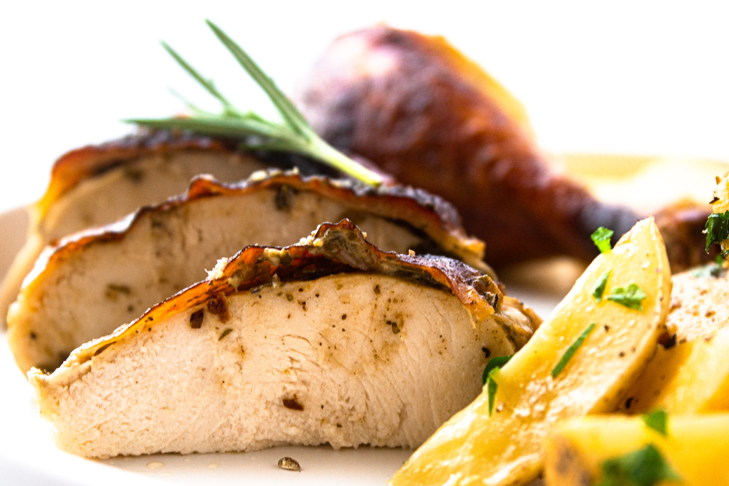 slices of roasted herbed chicken breast with a rosemary sprig on top and a chicken leg in the backgroudn with potatoes served on the side