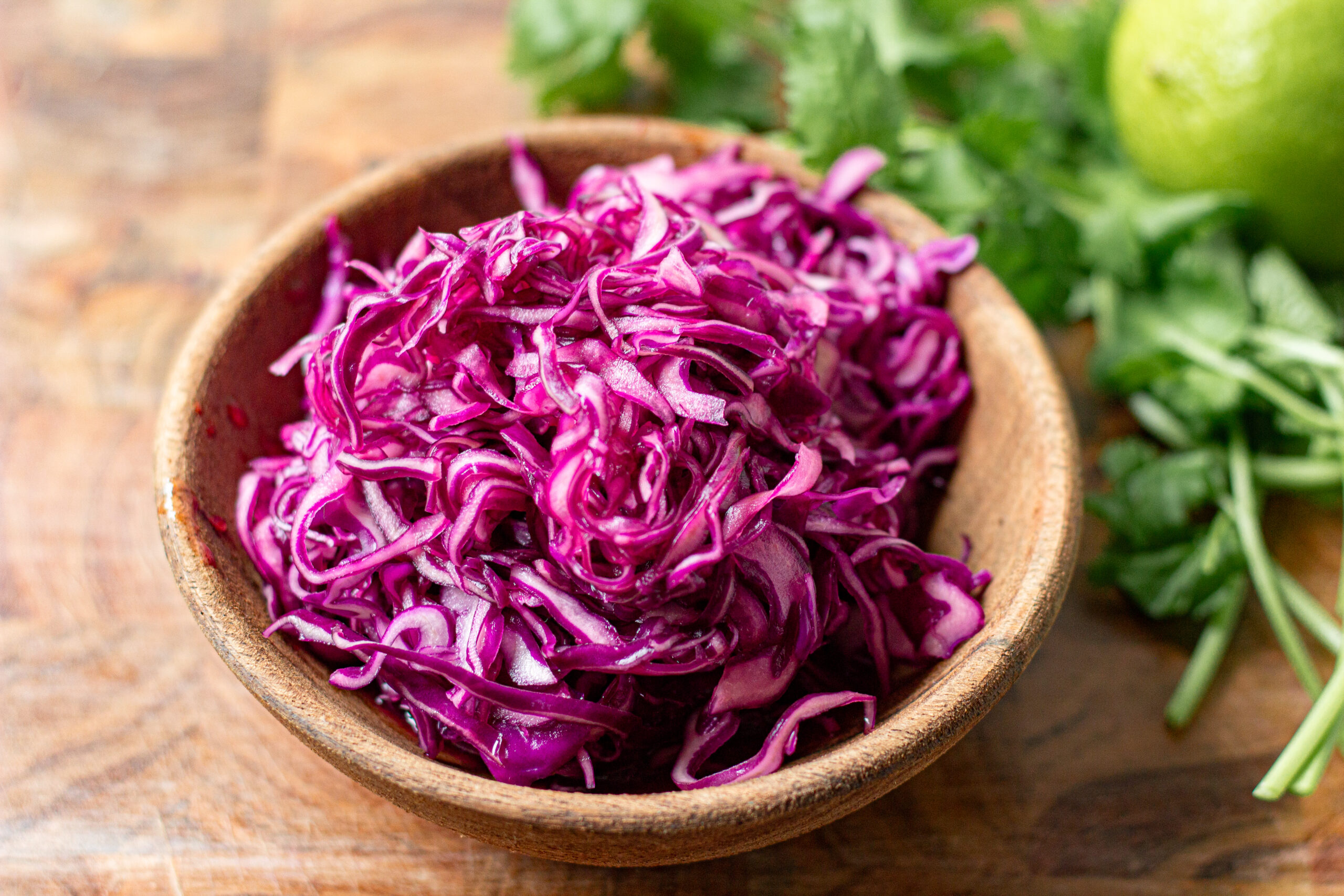 bright purple cabbage slaw in a wooden bowl with cilantro and lime in the background