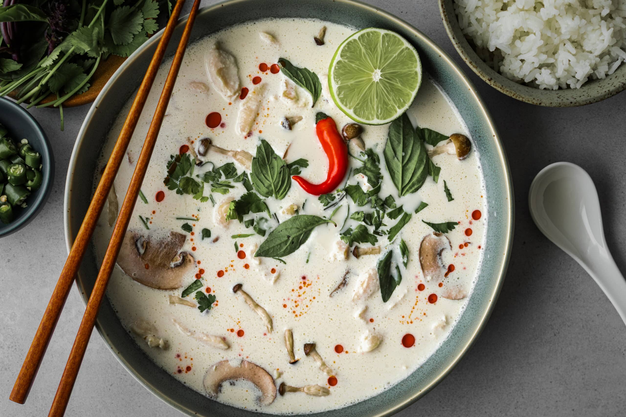 Tom Kha Gai soup in a wide ceramic bowl topped with fresh cilantro, Thai basil, a red chili pepper, sliced mushrooms, and a lime half, with chopsticks, a side of white rice, and fresh herbs in the background