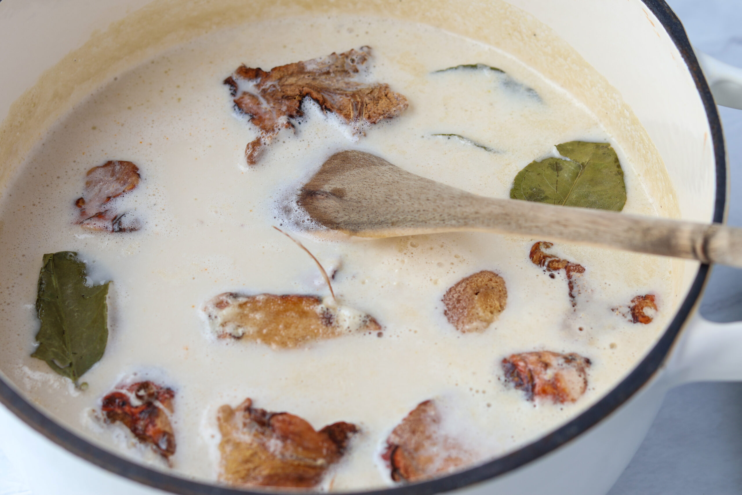 Coconut milk broth simmering in a Dutch oven with galangal, kaffir lime leaves, and dried chilies, stirred with a wooden spoon