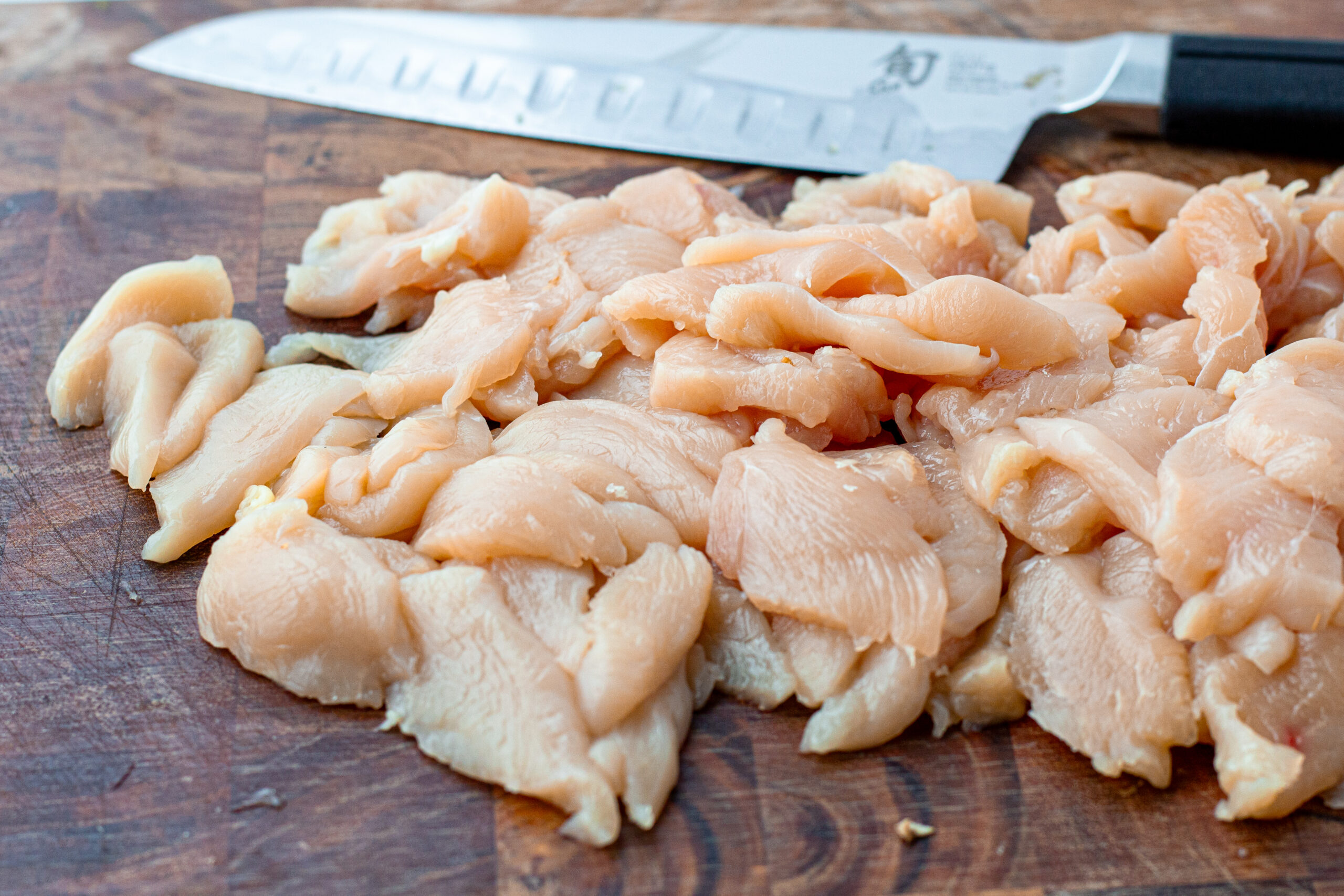 Thinly sliced raw chicken breast on a wooden cutting board with a chef's knife