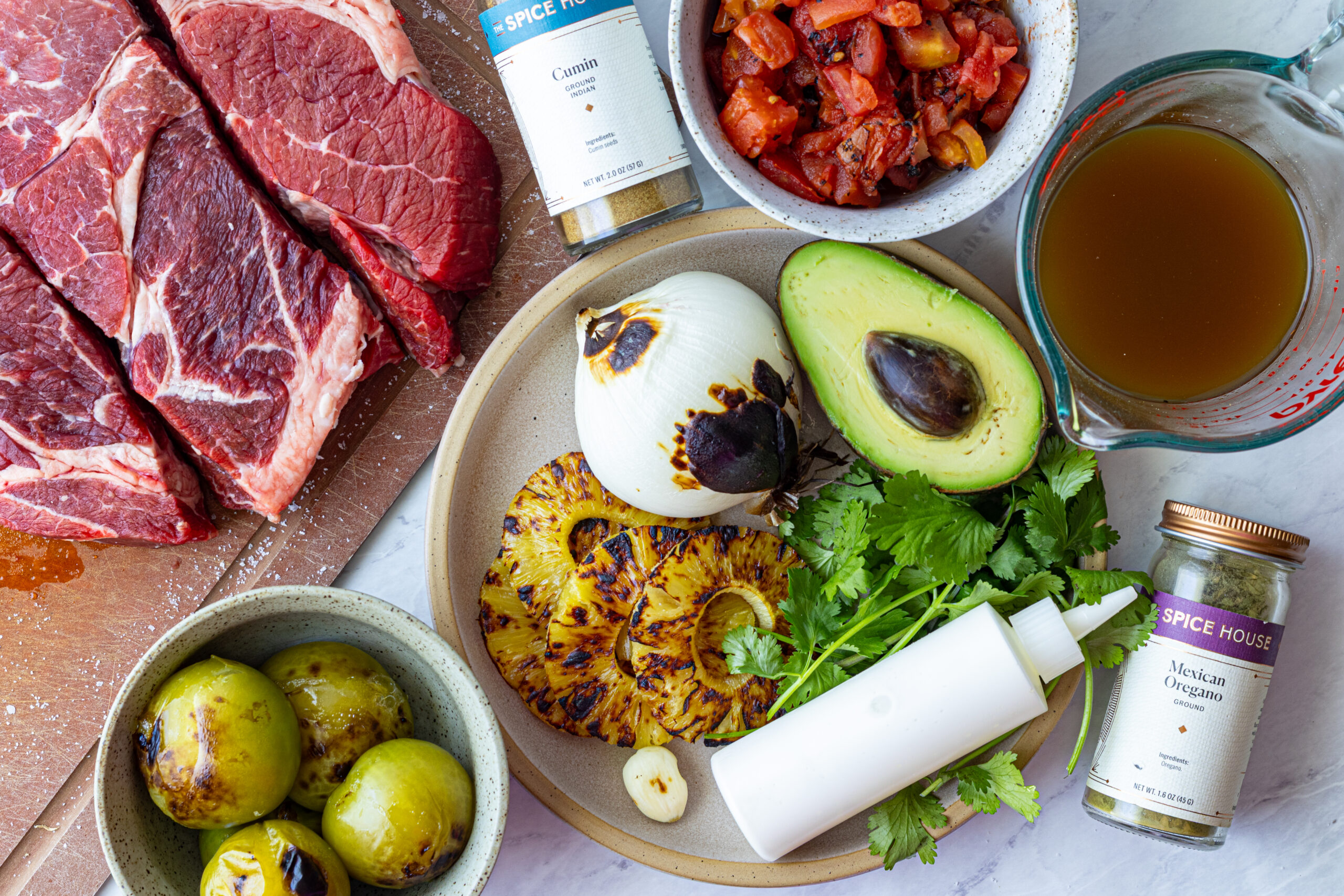 Beef tinga ingredients flat lay including chuck roast, roasted tomatillos, charred pineapple, avocado, cumin, Mexican oregano, cilantro, and beef broth