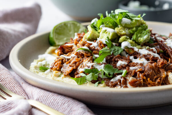 dish of beef tinga over creamy masa topped with smashed avocado, crema, cilantro, and a lime wedge, easy Mexican dinner recipe