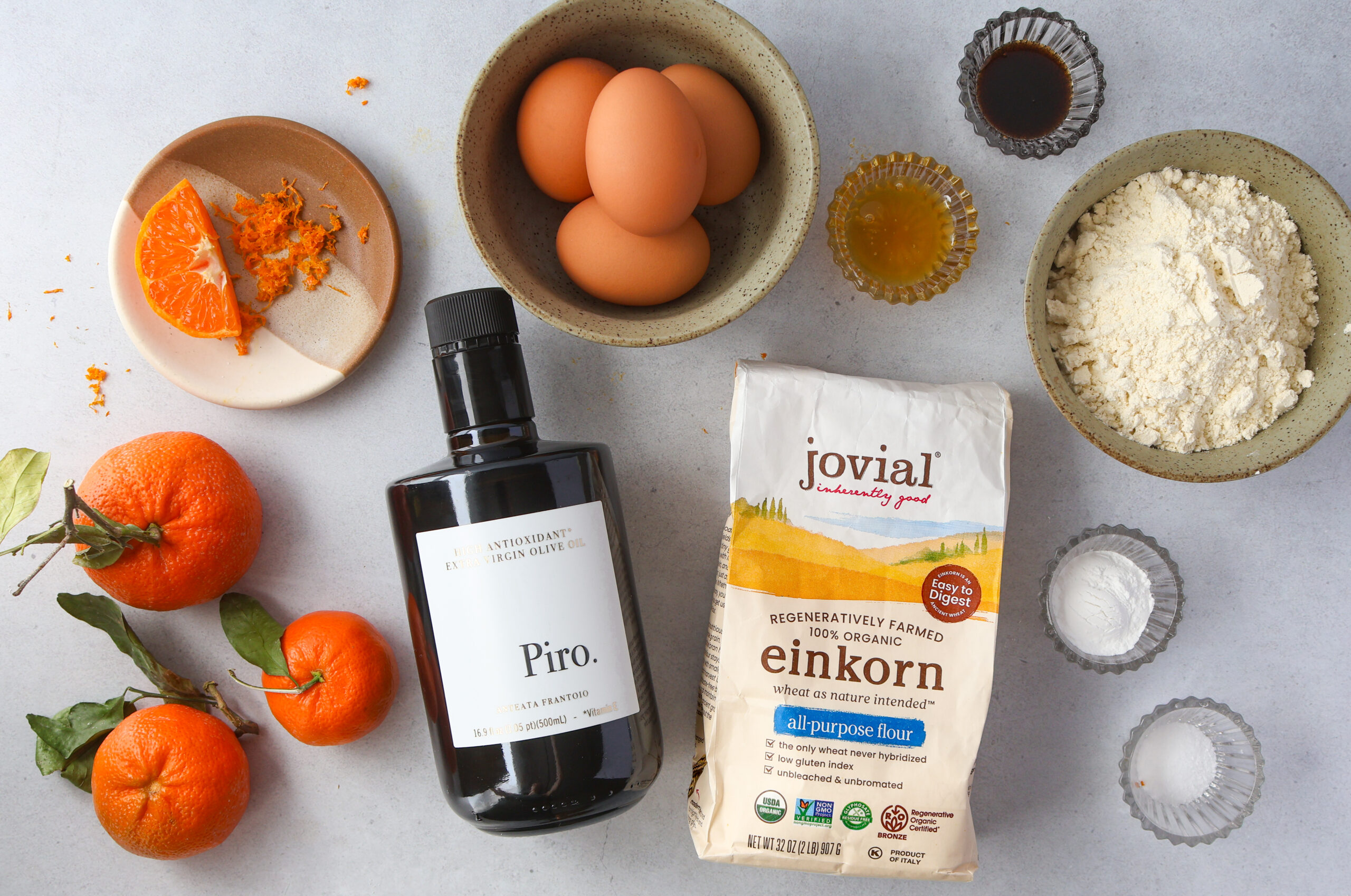Einkorn olive oil cake ingredients flat lay with Jovial einkorn flour, Piro extra virgin olive oil, eggs, clementines, honey, vanilla, baking powder, and salt