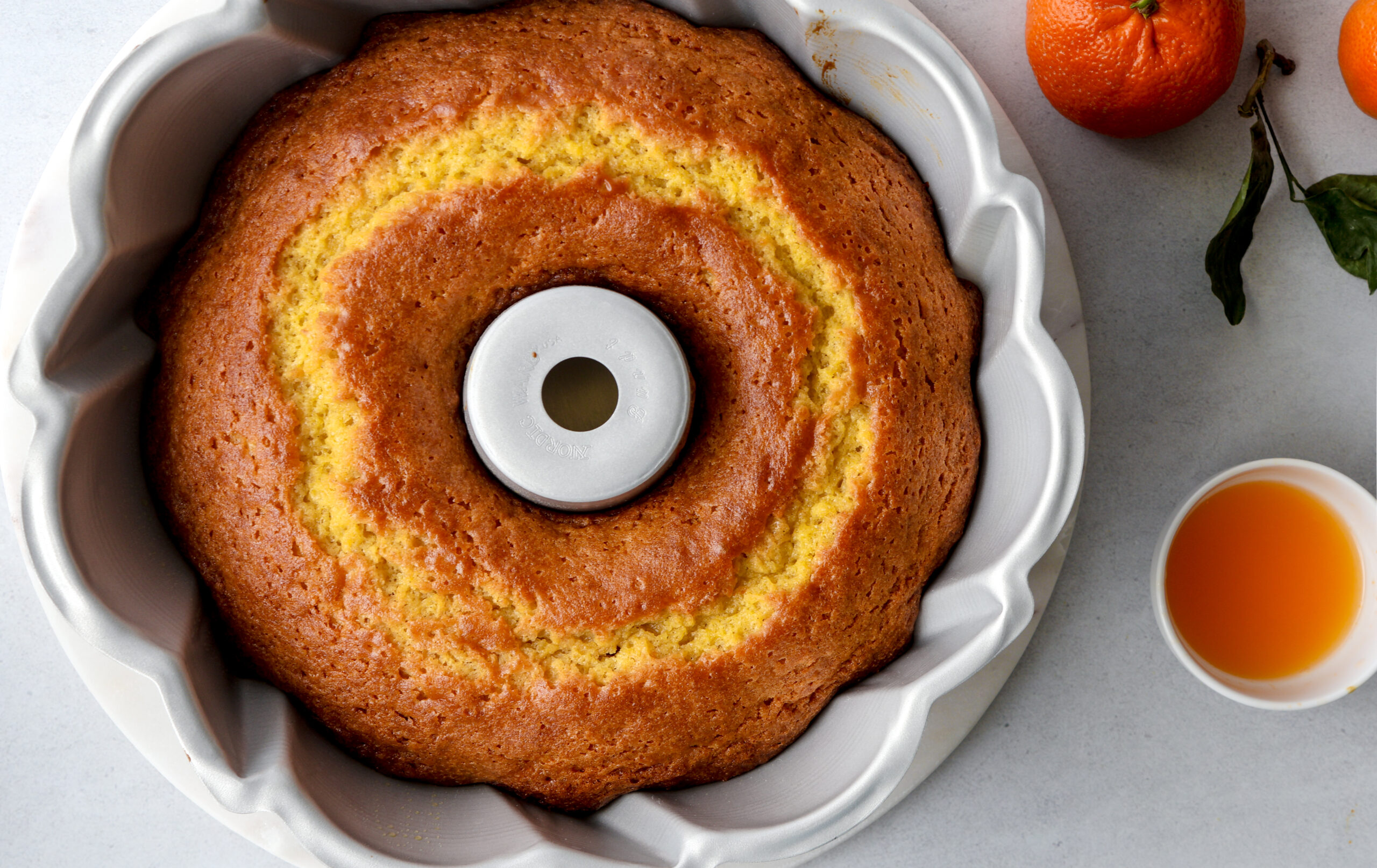Einkorn olive oil cake fresh from the oven in a white bundt pan, overhead view with clementines and small bowl of honey alongside