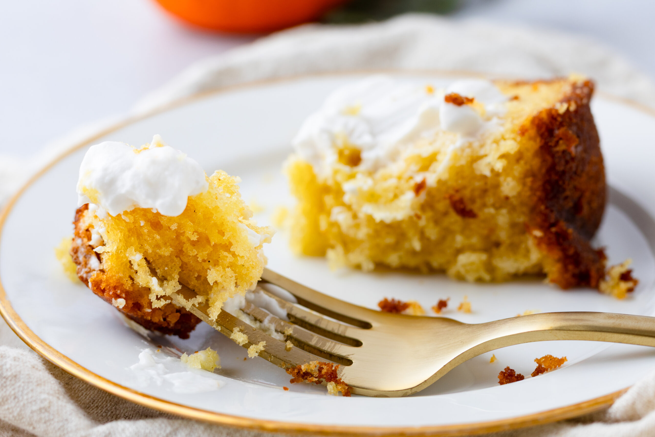 
Close up of a bite of einkorn olive oil cake on a gold fork showing a moist golden crumb, slice with whipped cream on a white gold-rimmed plate