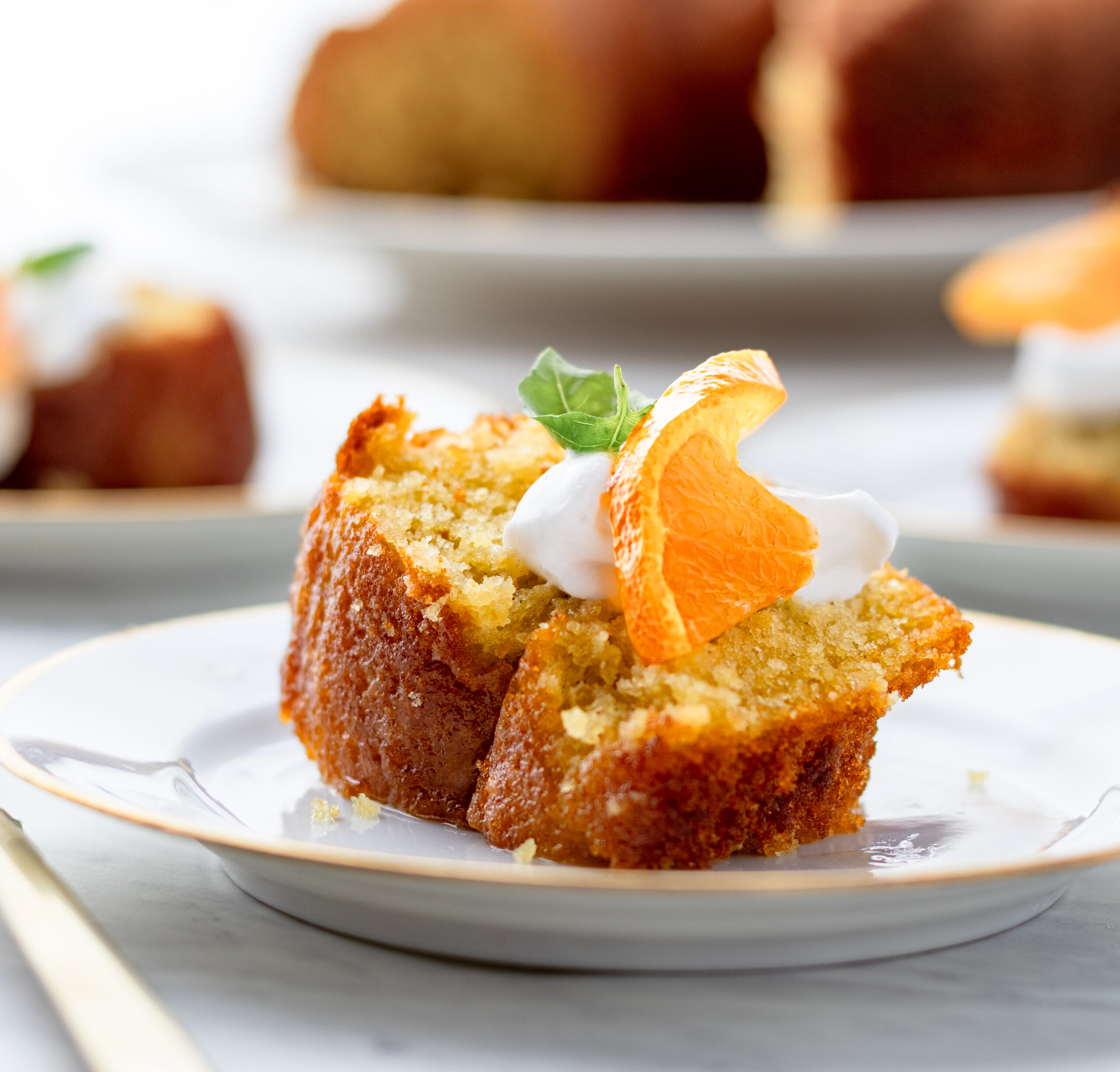 Slice of einkorn olive oil cake topped with whipped cream and a fresh clementine wedge on a white plate, whole cake blurred in background