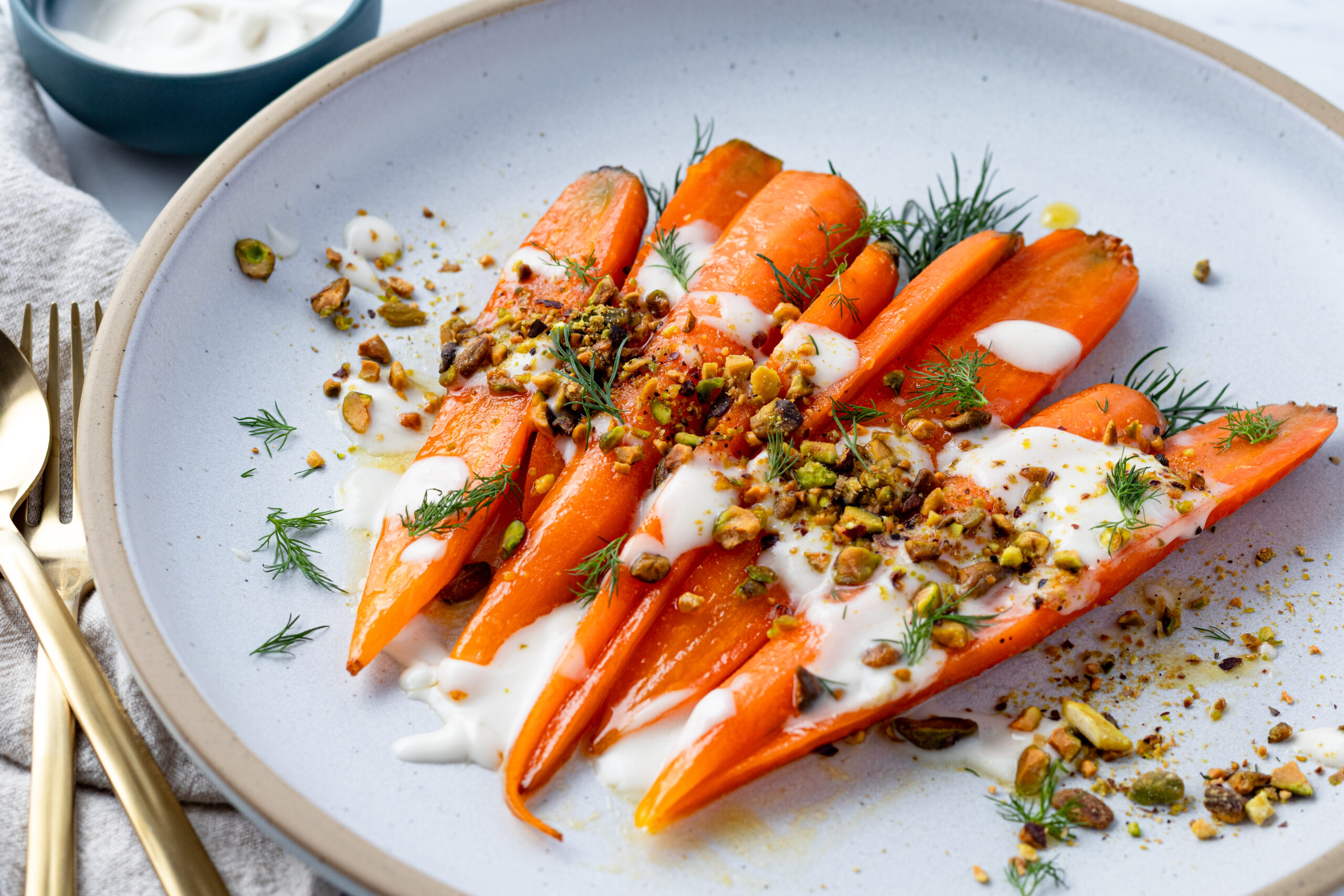 Honey glazed whole carrots served on a white ceramic plate with honey yogurt sauce, crushed toasted pistachios, and fresh dill garnish, with gold fork and knife on a linen napkin