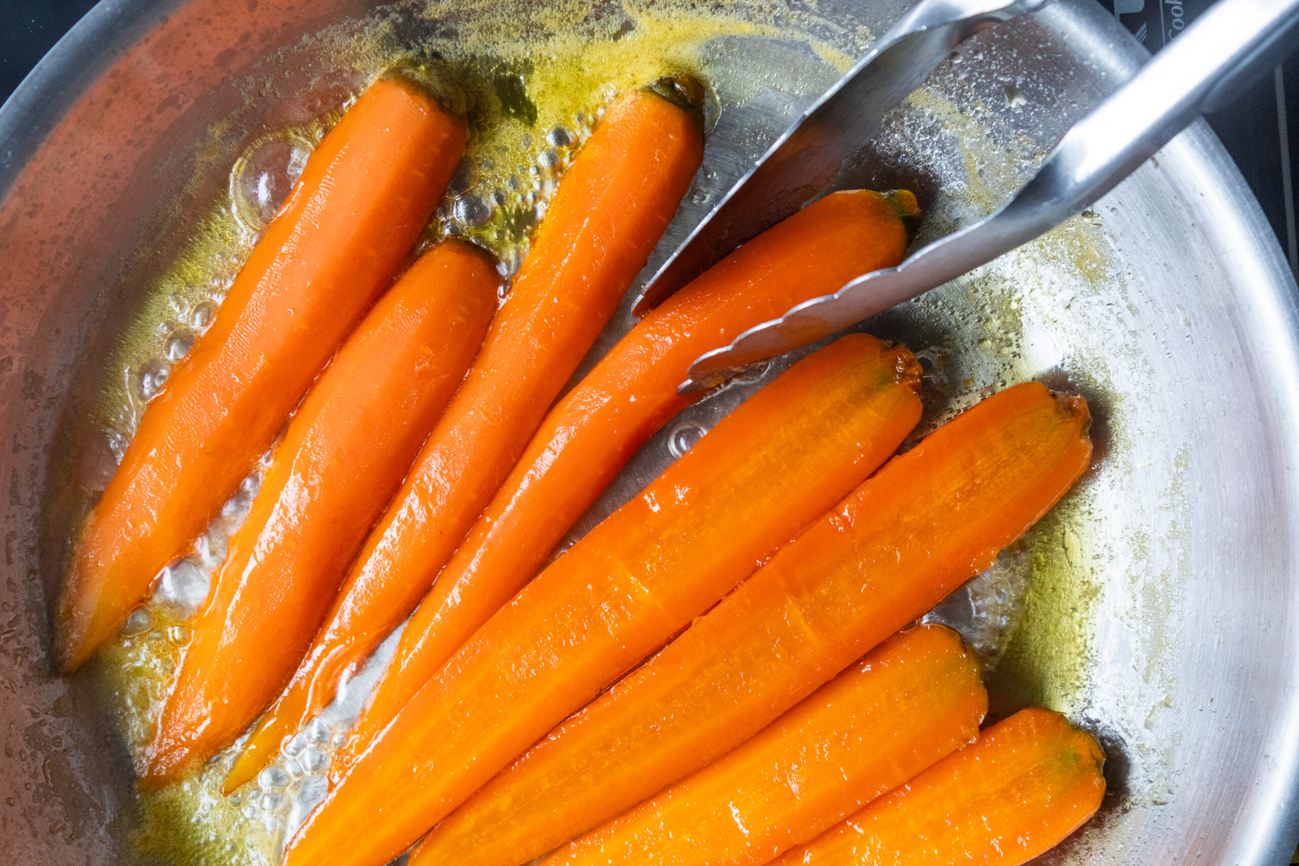 halfted carrots being glazed in a bubbling honey butter sauce in a stainless steel pan with tongs, easy stovetop carrot recipe