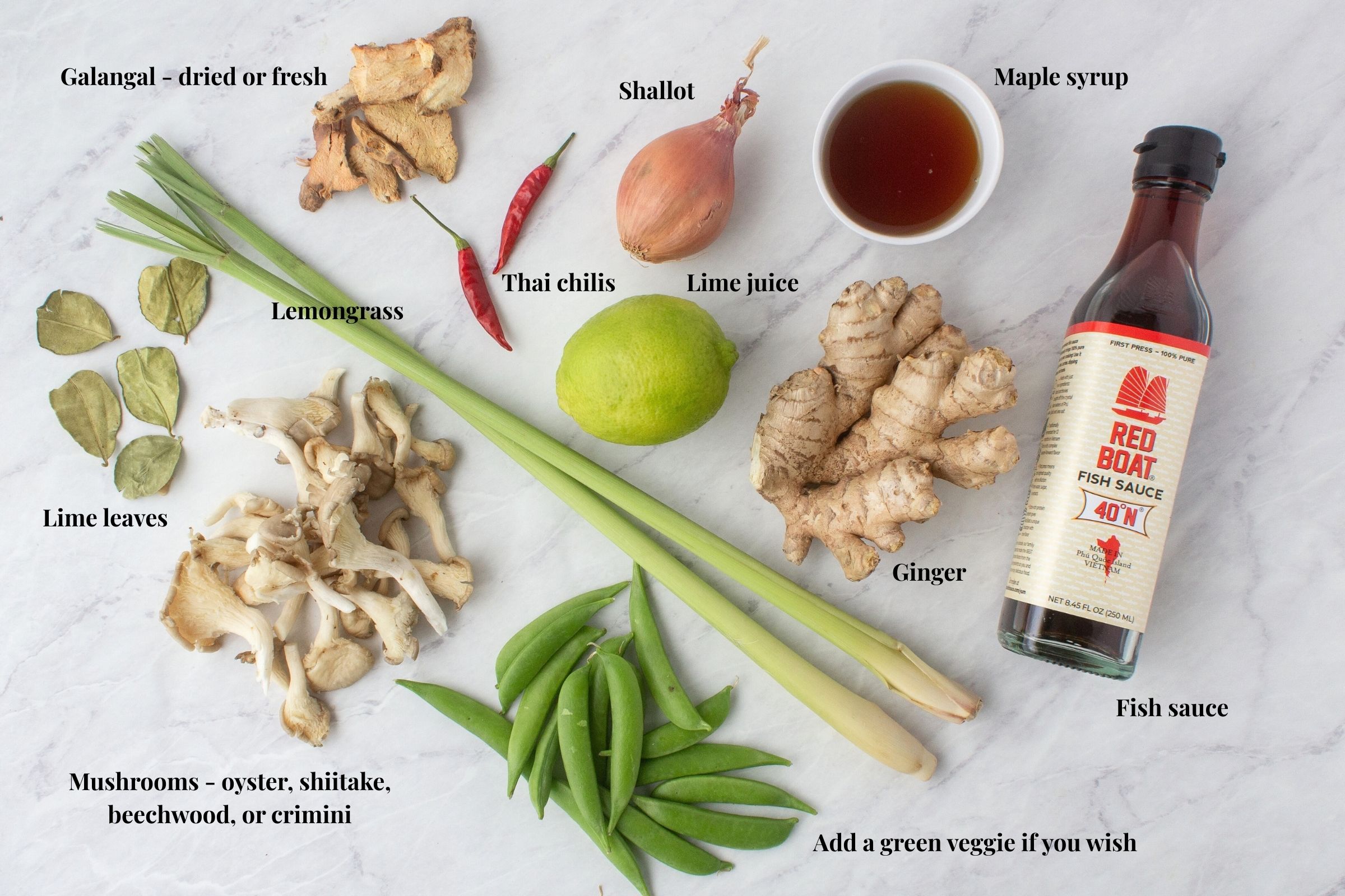 Tom Kha Gai ingredients flat lay on marble including galangal, lemongrass, kaffir lime leaves, Thai chilies, mushrooms, ginger, shallot, lime, fish sauce, and snap peas