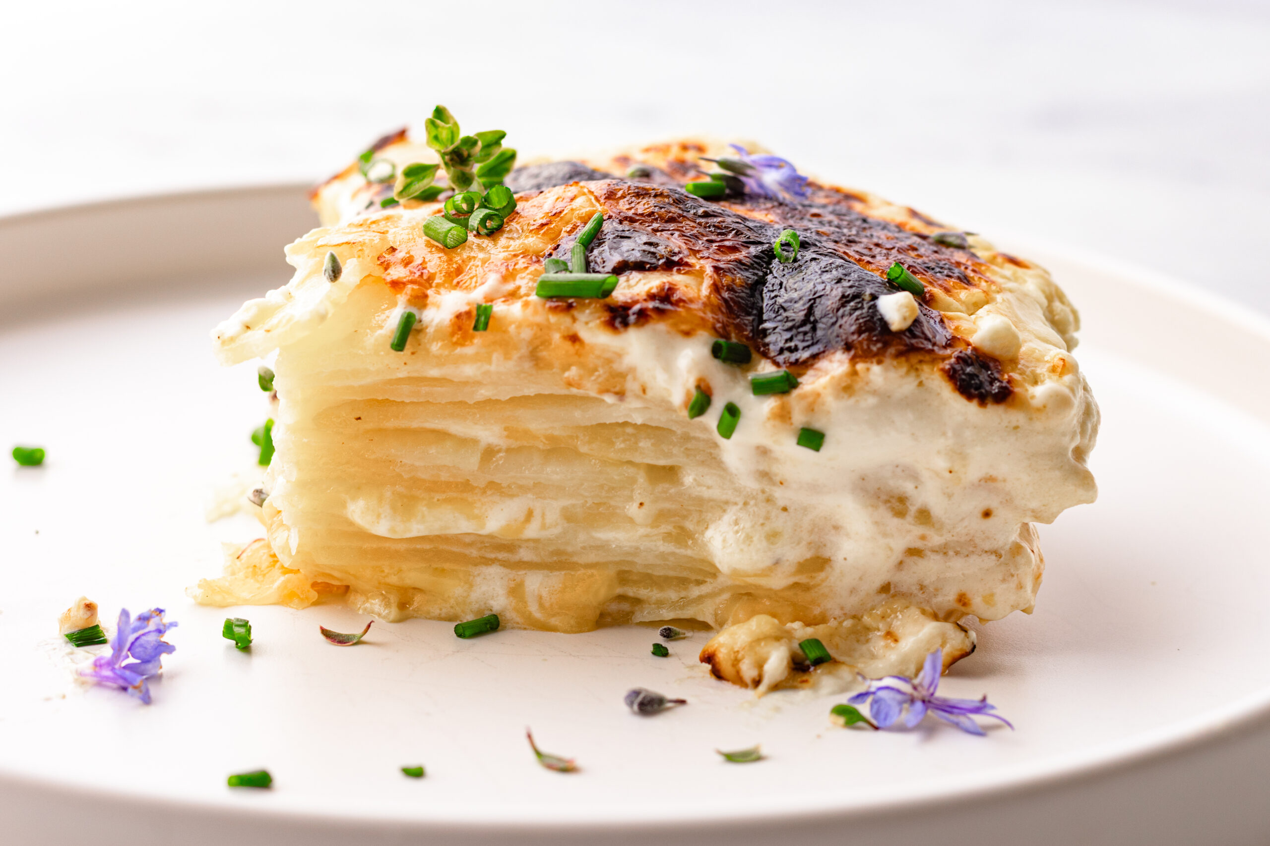 A slice of au gratin potatoes on a white plate showing creamy layered russet potatoes with a golden browned crust, garnished with chives and edible flowers