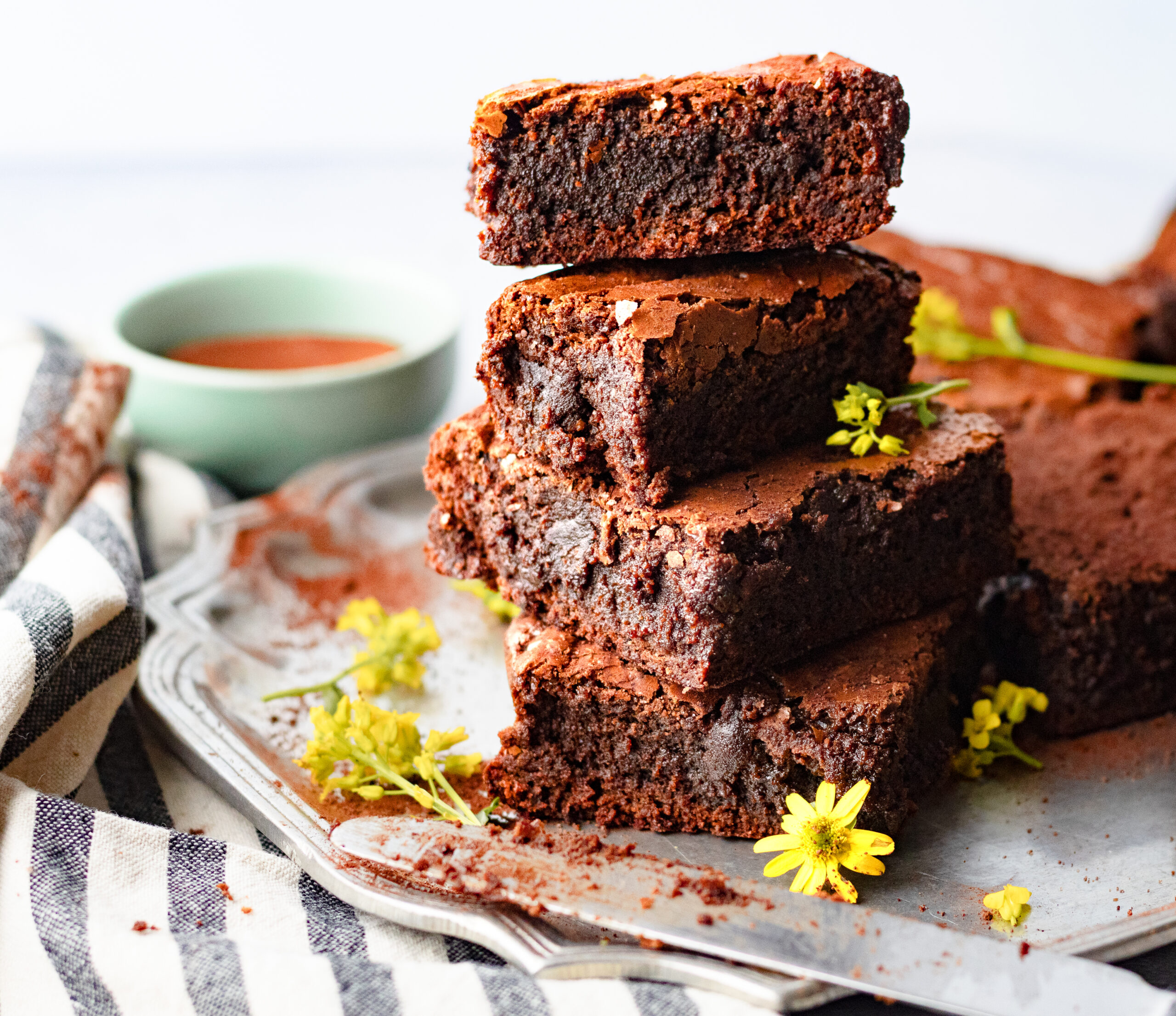 Stack of four fudgy einkorn brownies on a vintage silver tray showing the dense gooey interior with yellow flower garnish