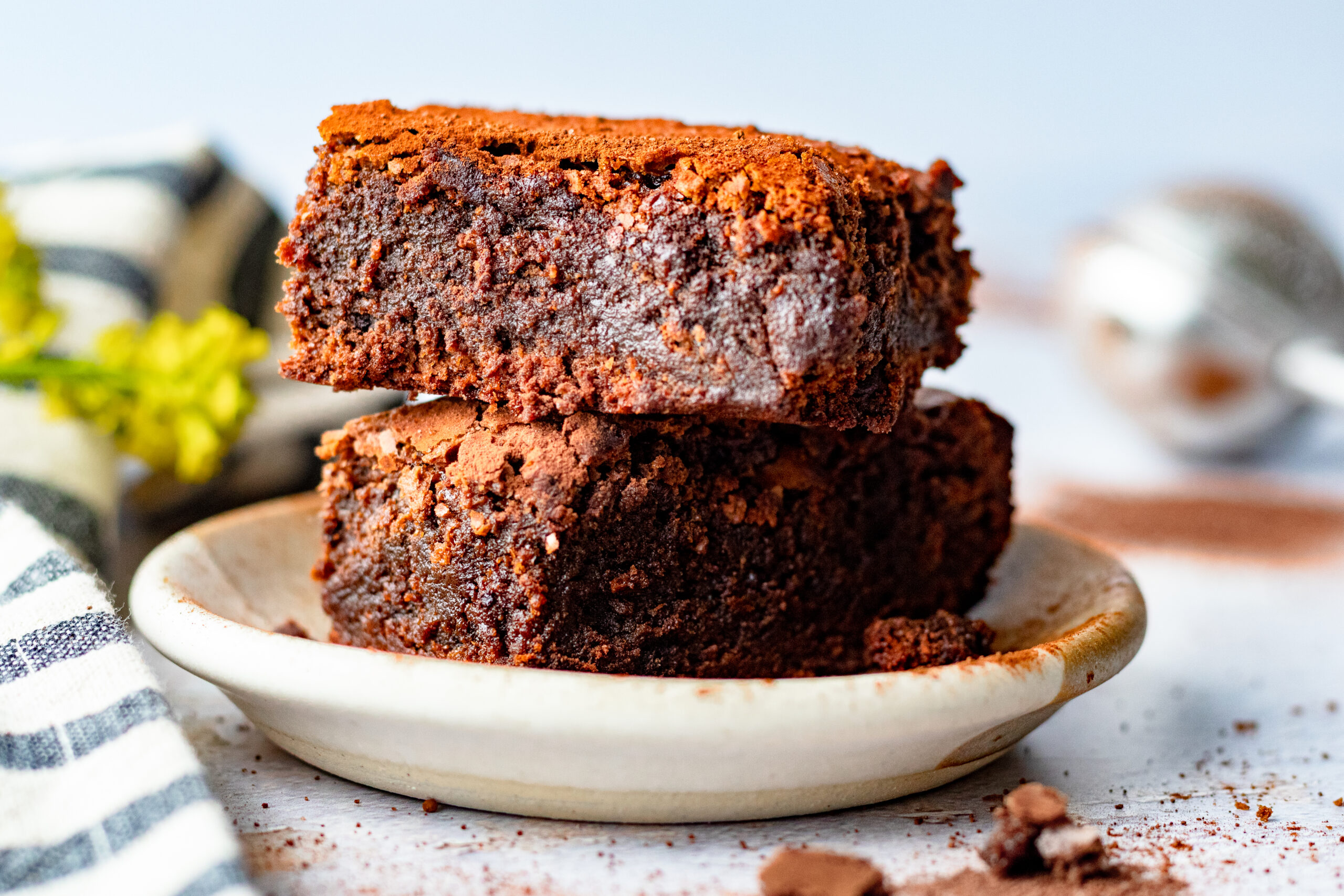 Two brownies stacked on a ceramic plate dusted with cocoa powder with yellow flowers and striped linen in the background
