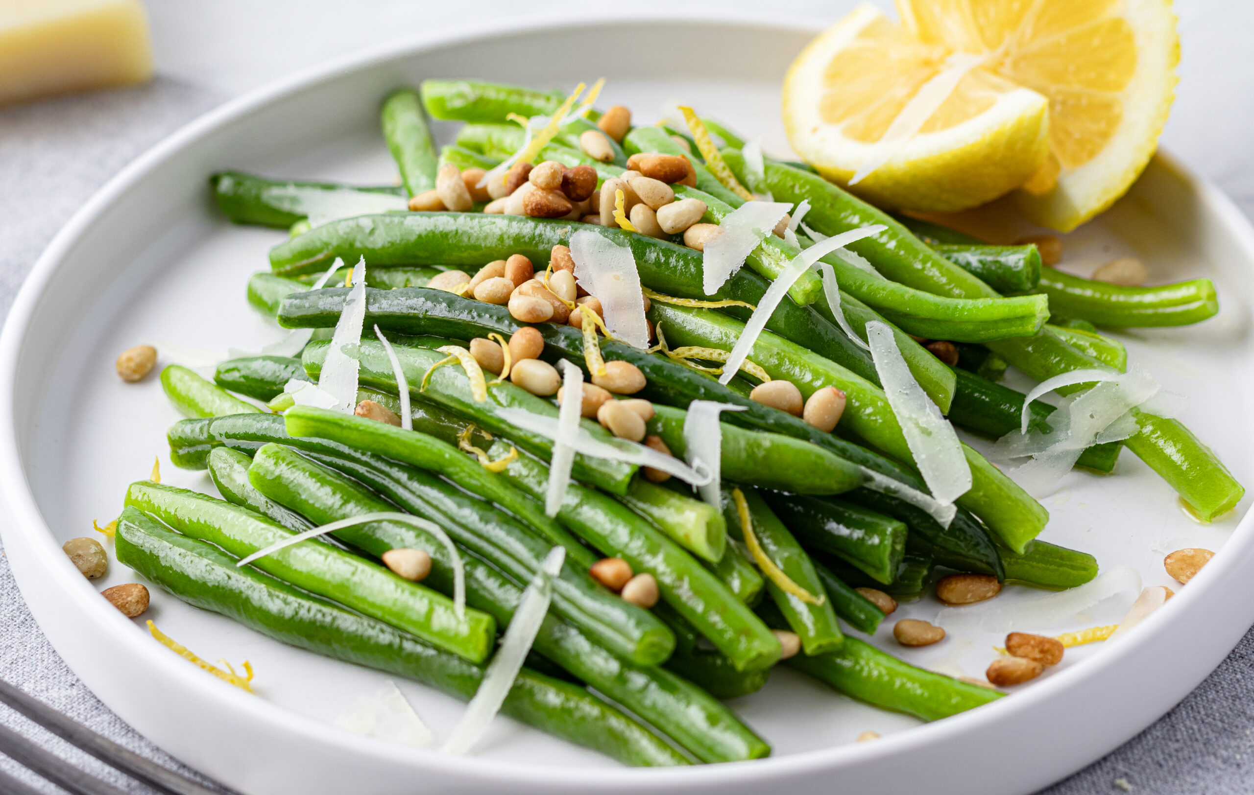 Perfectly blanched green beans on a white plate topped with toasted pine nuts, shaved Pecorino Romano, and fresh lemon zest with a halved lemon in the background