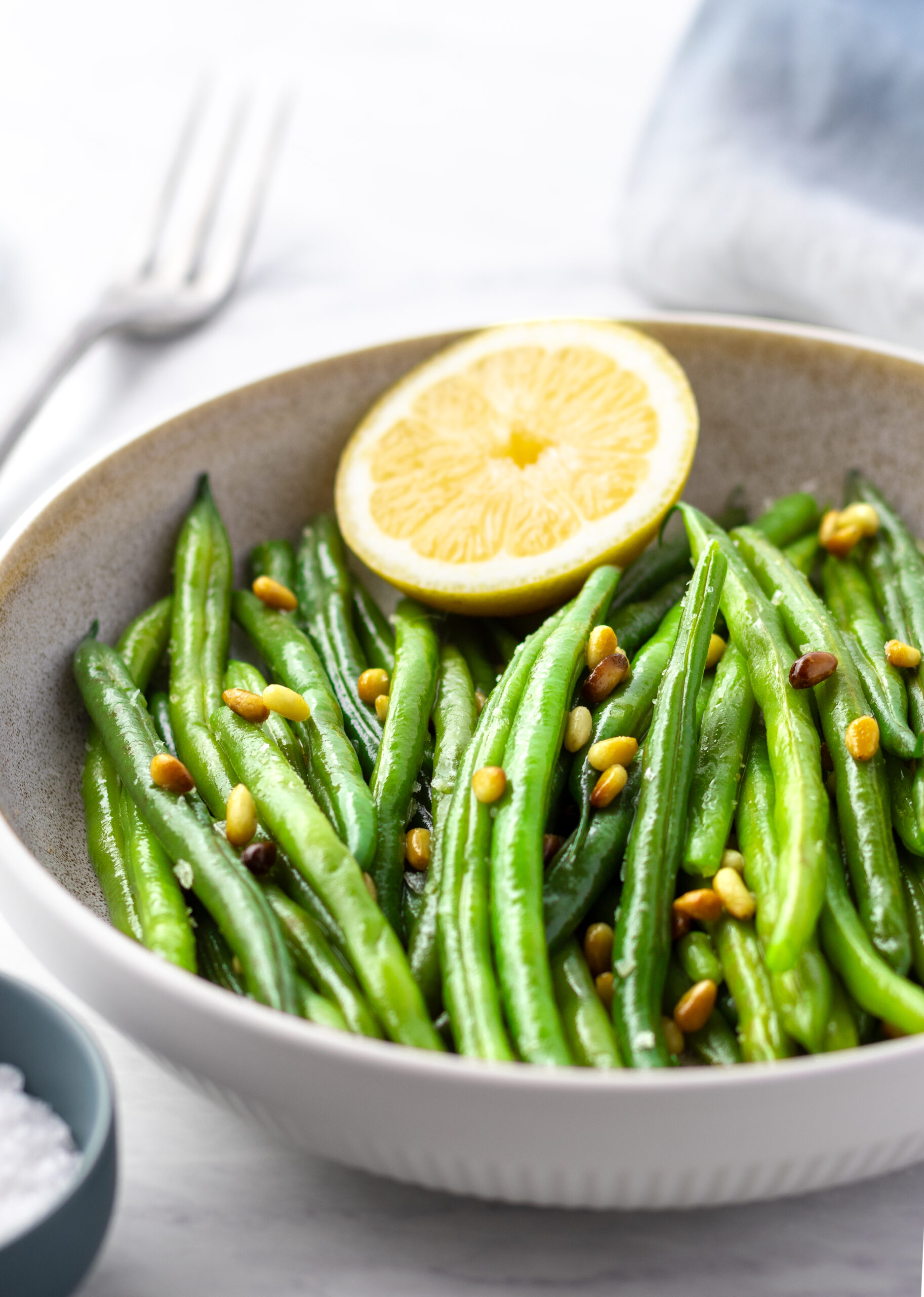 Tender blanched green beans in a white bowl topped with toasted pine nuts and a halved lemon with a small dish of flaky sea salt on the side