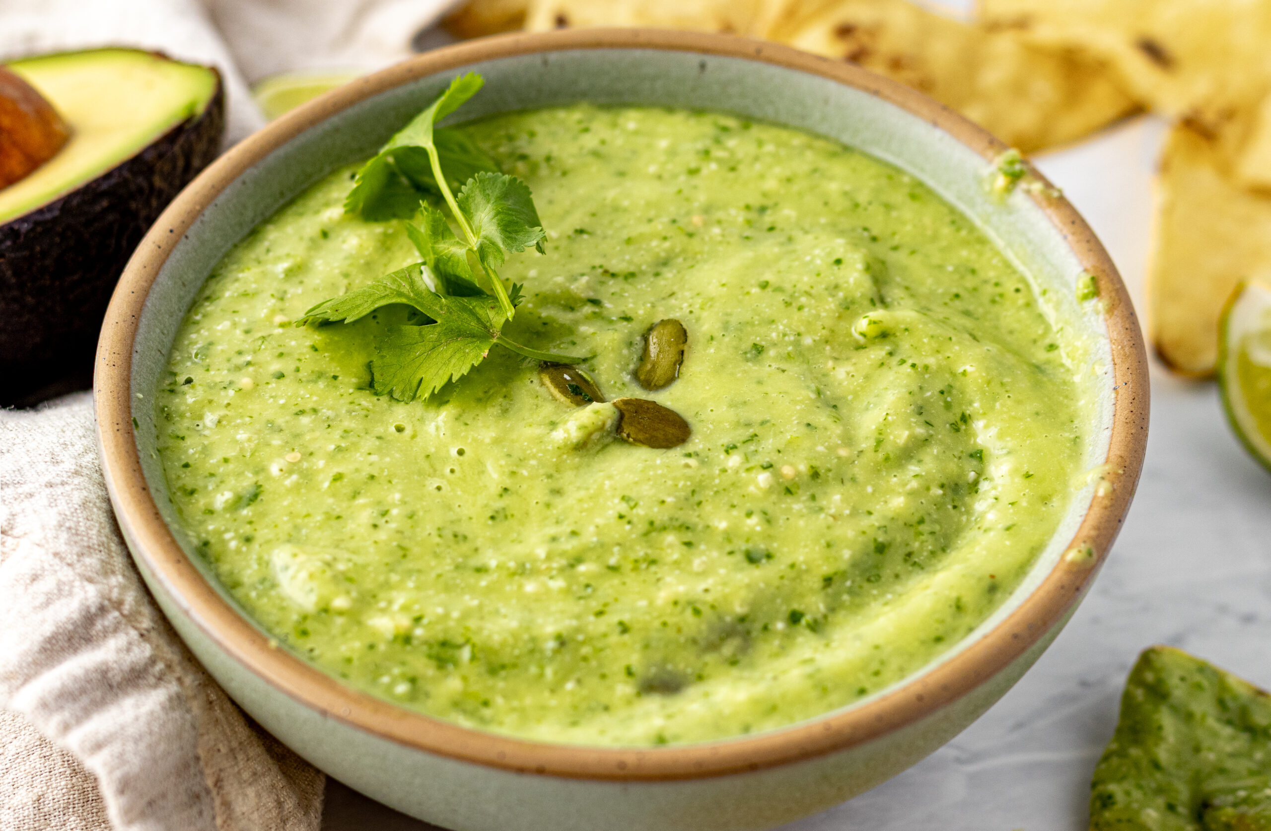 Close-up of creamy pepita avocado salsa in a ceramic bowl garnished with fresh cilantro and toasted pepitas, with avocado and tortilla chips in the background