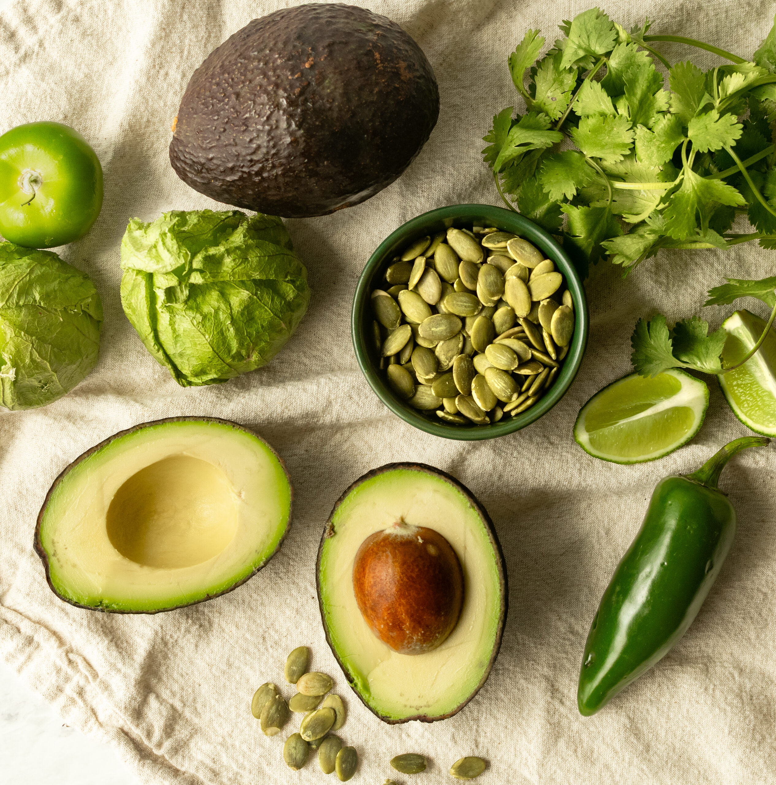 Flat lay of pepita avocado salsa ingredients including whole and halved avocados, tomatillos, pepitas, jalapeño, fresh cilantro, and lime wedges on a linen cloth