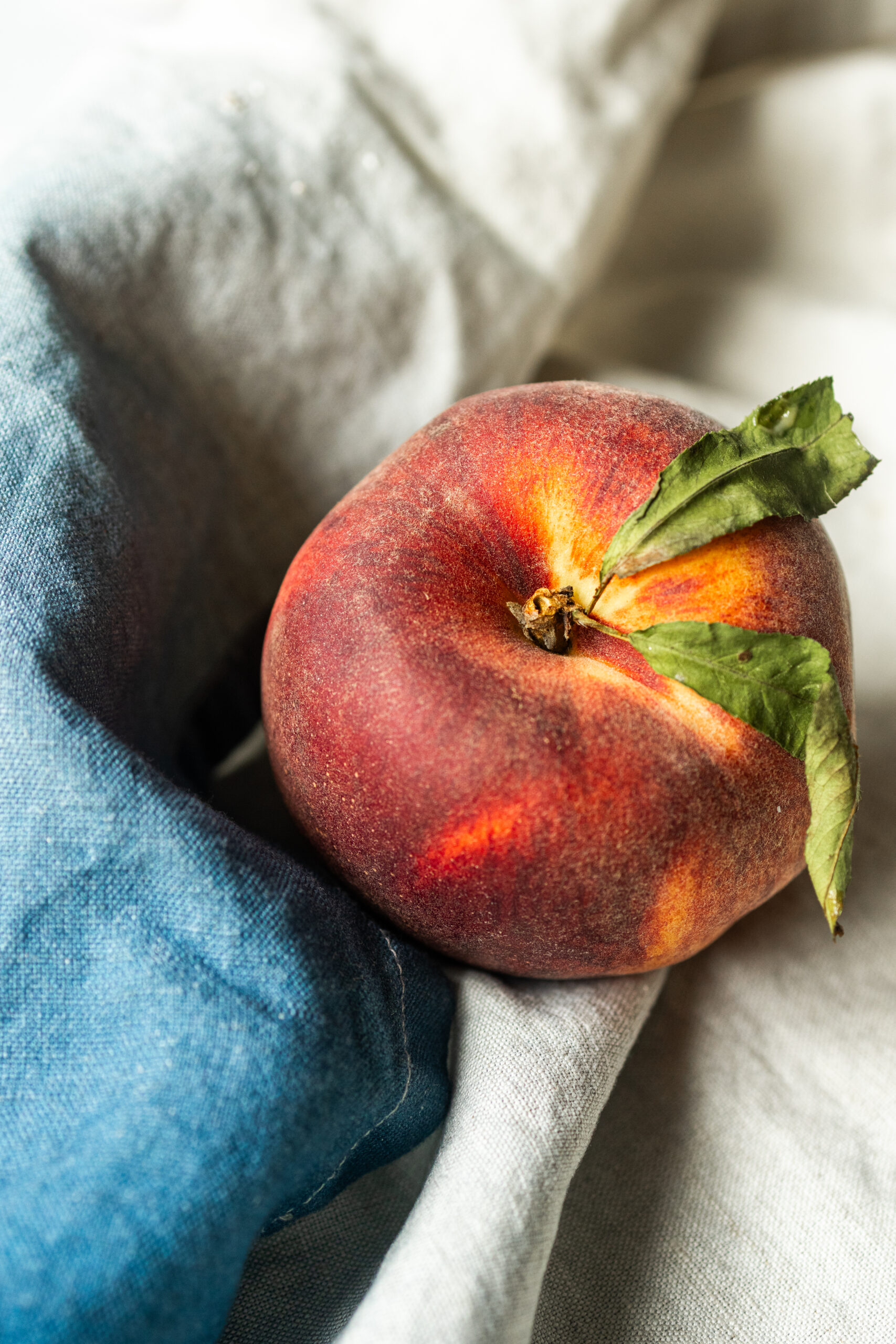 A ripe red and orange peach with green leaves resting on linen and denim fabric