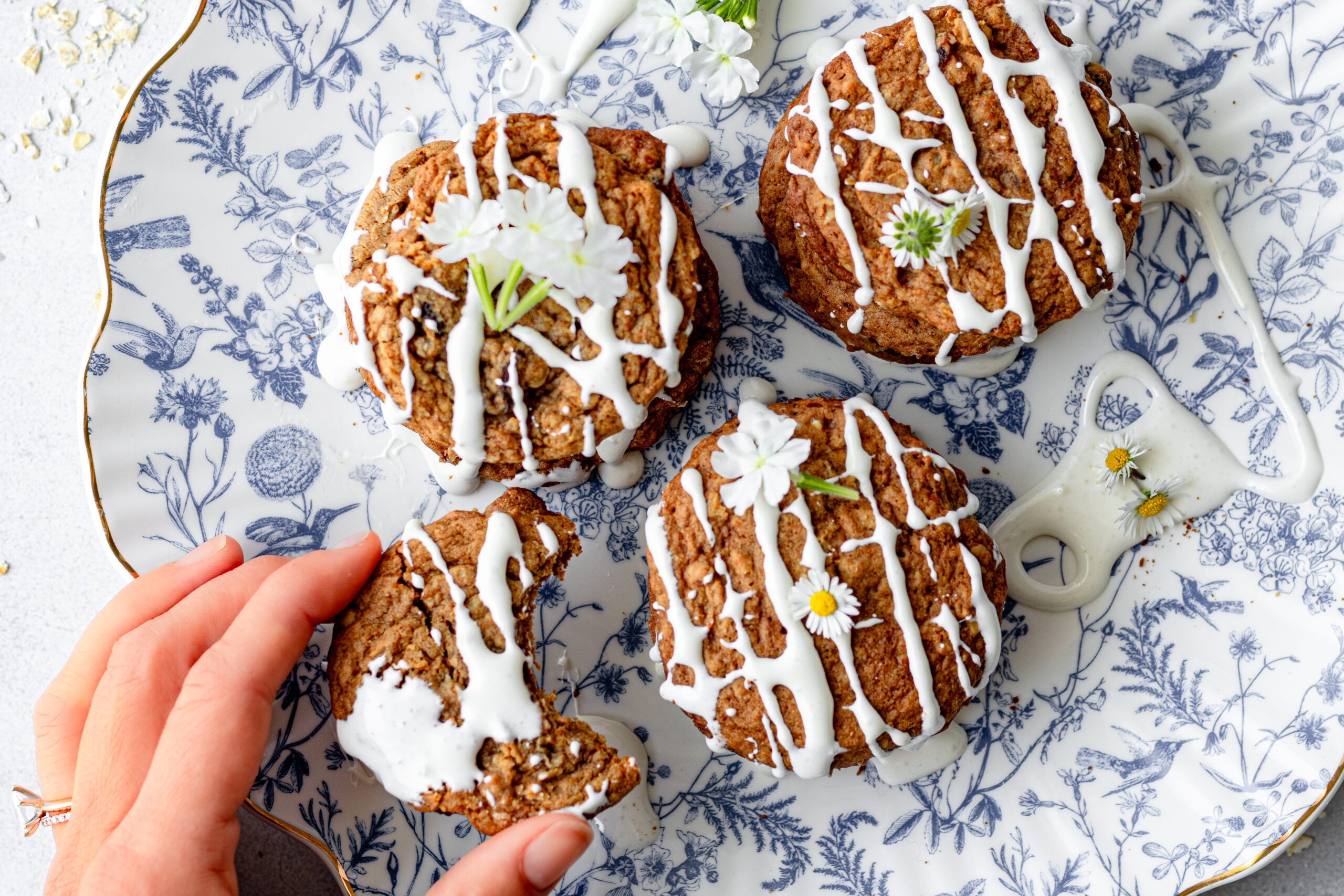 Overhead view of iced einkorn oatmeal raisin cookies on a blue and white toile plate garnished with chamomile flowers and a hand reaching in