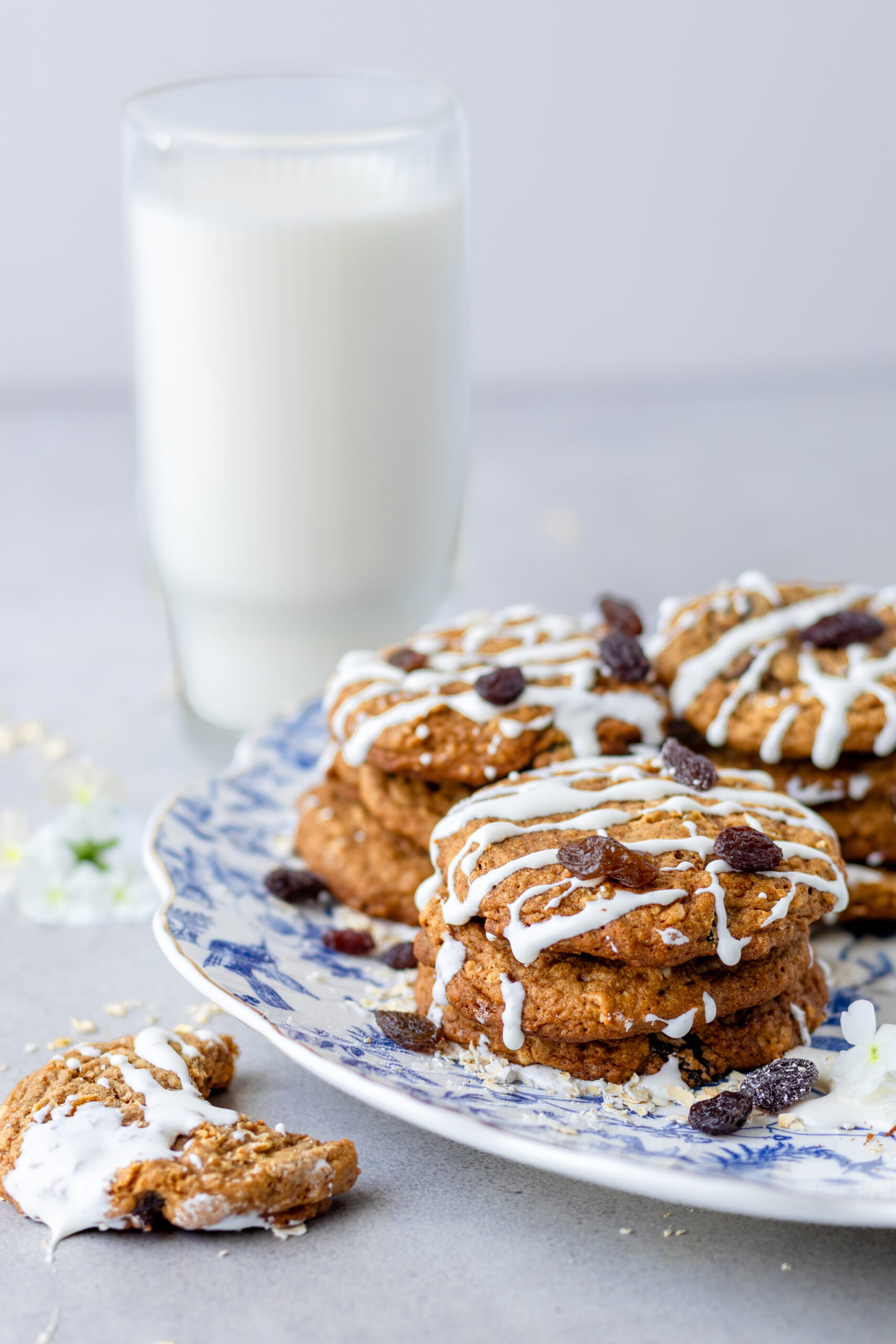 Iced einkorn oatmeal raisin cookies stacked on a blue and white toile plate with raisins scattered on top and a glass of milk in the background