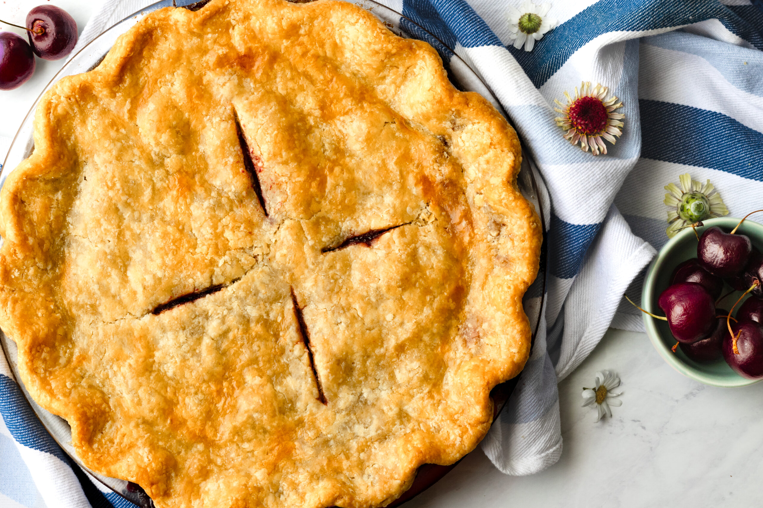 Golden baked einkorn cherry pie with crimped edges and four vent slits, topped with fresh cherries and small dried flowers on a blue striped linen