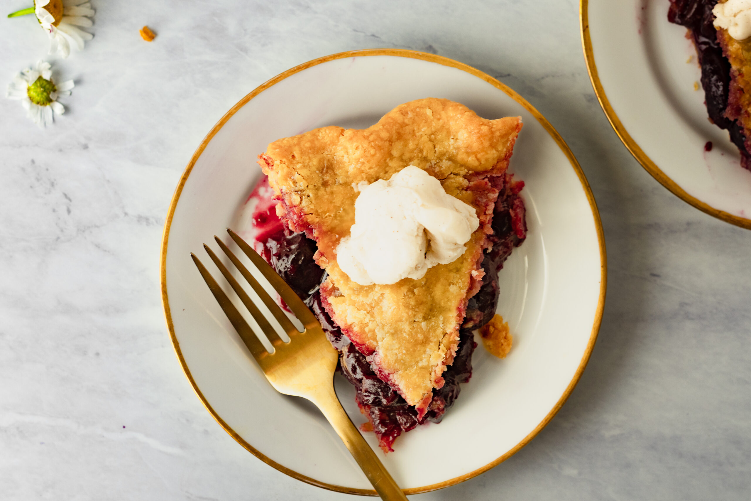 Overhead slice of einkorn cherry pie topped with a scoop of vanilla ice cream on a white and gold plate with a gold fork, with a second slice and daisies in the background