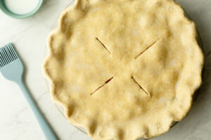 Unbaked einkorn double crust pie with crimped edges and four vent slits cut in an X pattern, with a pastry brush and small bowl of milk nearby