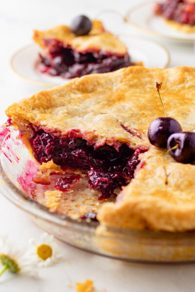 Sliced einkorn cherry pie in a glass pie dish showing the dark sweet cherry filling, with fresh cherries resting on the crust and a plated slice in the background
