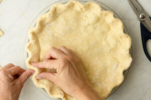Hands crimping the edges of einkorn pie dough in a glass pie dish with kitchen scissors nearby