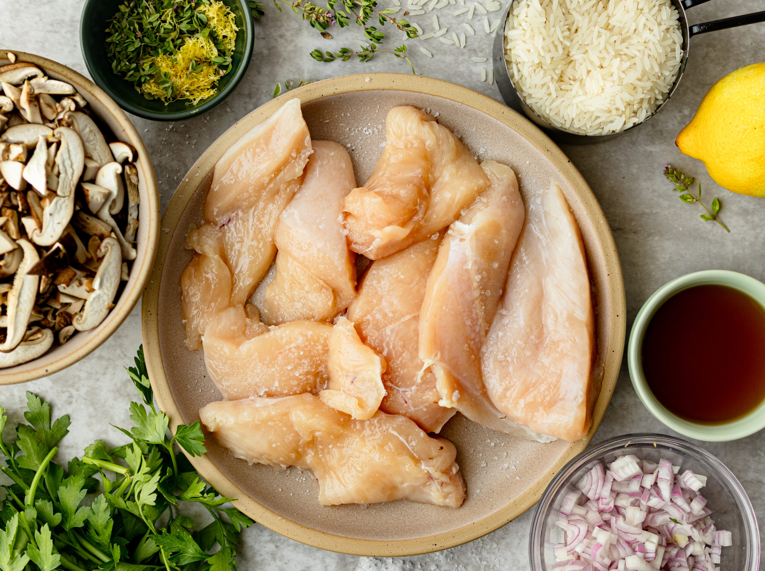 Flat lay of one pan chicken and mushroom rice ingredients including raw chicken tenders, sliced crimini and shiitake mushrooms, jasmine rice, lemon zest, fresh thyme, diced shallot, and marsala wine on a gray surface.