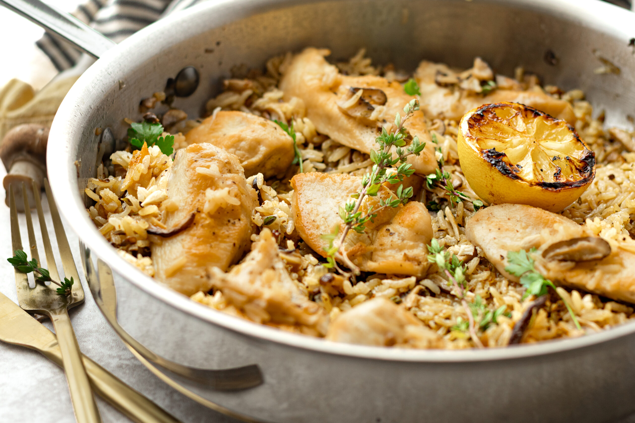 Angled shot of one pan chicken and mushroom rice in a stainless skillet garnished with fresh thyme, parsley, and a broiled lemon half, with gold forks resting alongside on a yellow linen.