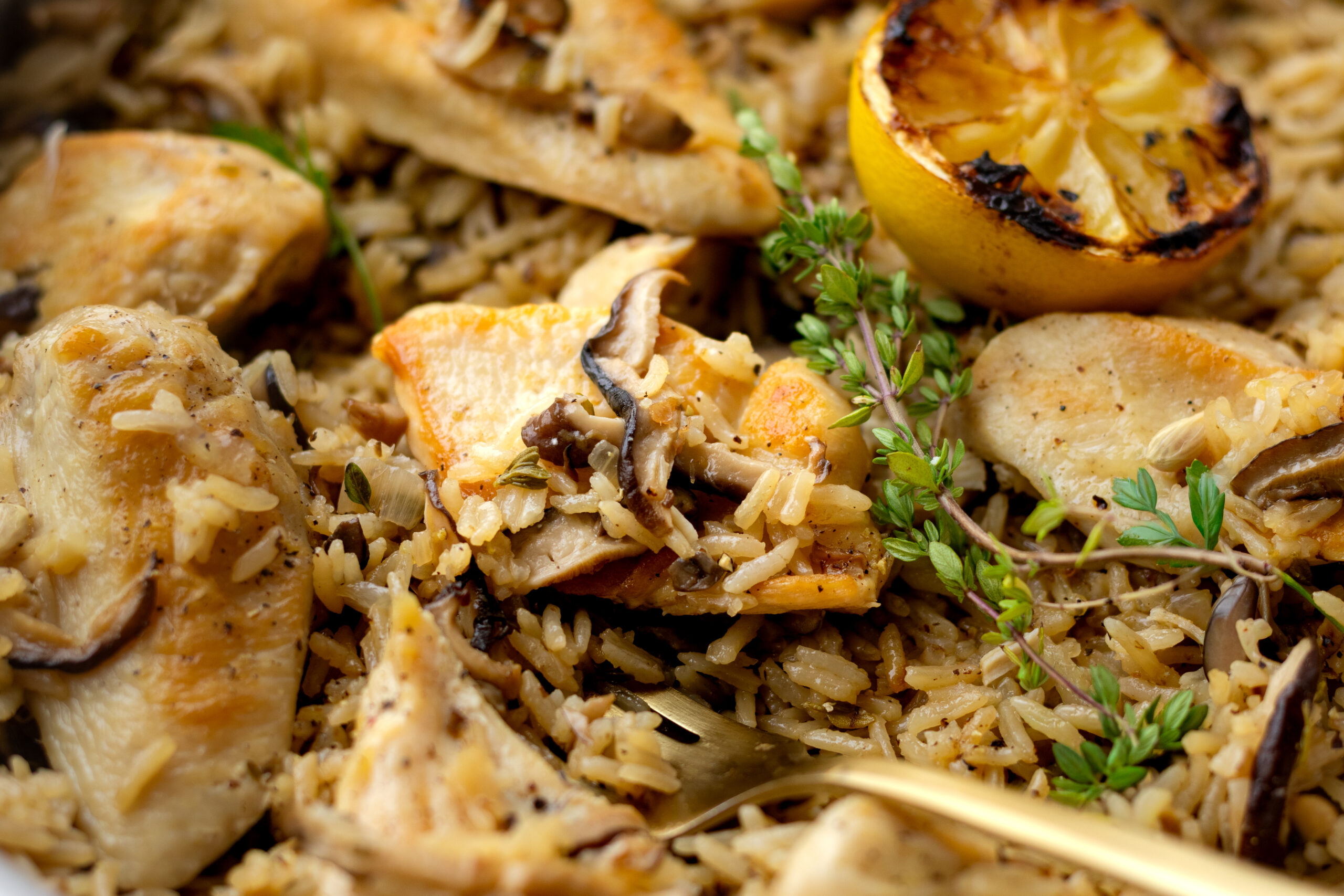 Close up of one pan chicken and mushroom rice showing tender chicken pieces nestled into savory jasmine rice with shiitake mushrooms, fresh thyme sprigs, and a charred lemon half in the background.